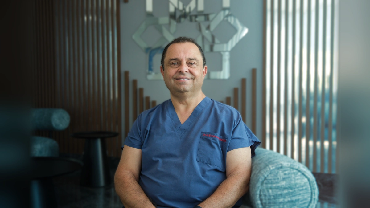 Prof. Dr. Mustafa Ercüment Önder, mavi sağlık çalışanı formasıyla otururken kameraya gülümsüyor.