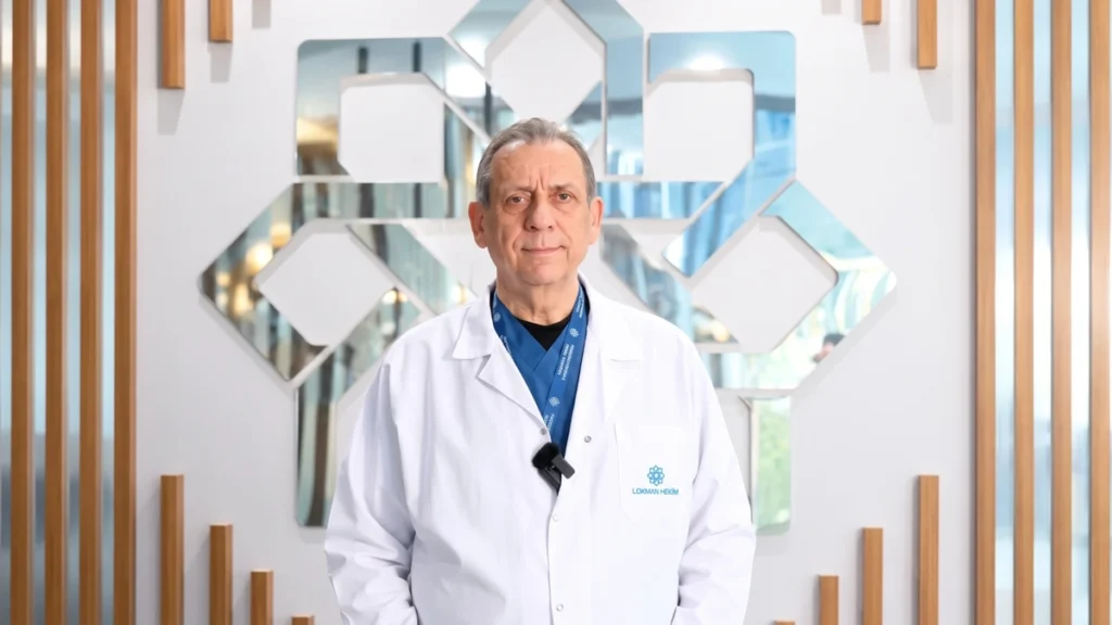 Prof. Dr. Atilla Stephan Ataç, beyaz hekim önlüğüyle ahşap çıtalı ve aynalı bir duvar panosunun önünde kameraya bakıyor.